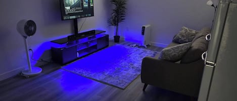 A living room with a flat-screen TV, a standing fan, a potted plant, a sofa, and a window with blinds.