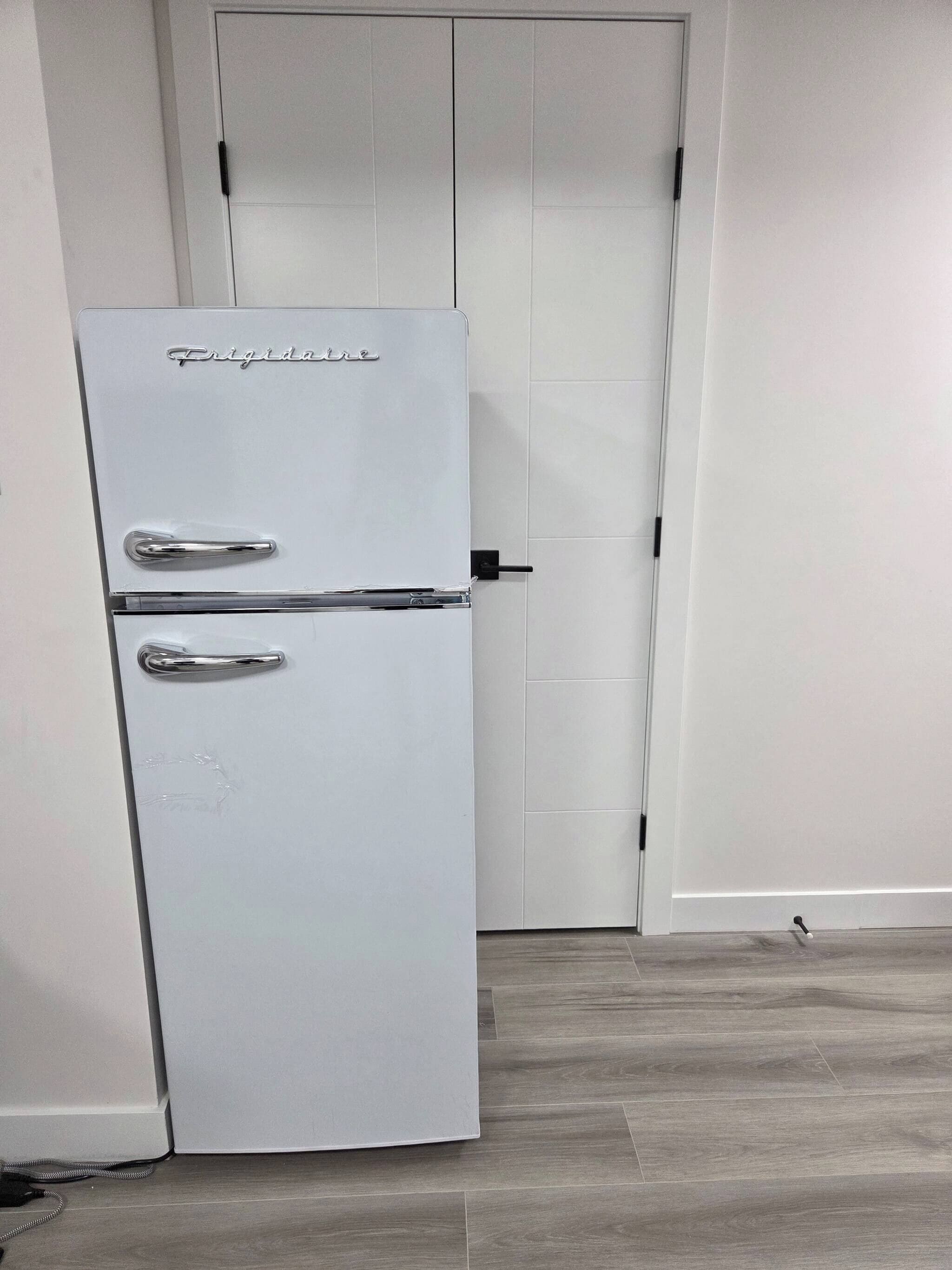 Un refrigerador blanco con un manija y un compartimento congelador, ubicado en una habitación con piso de madera y una puerta blanca al fondo.