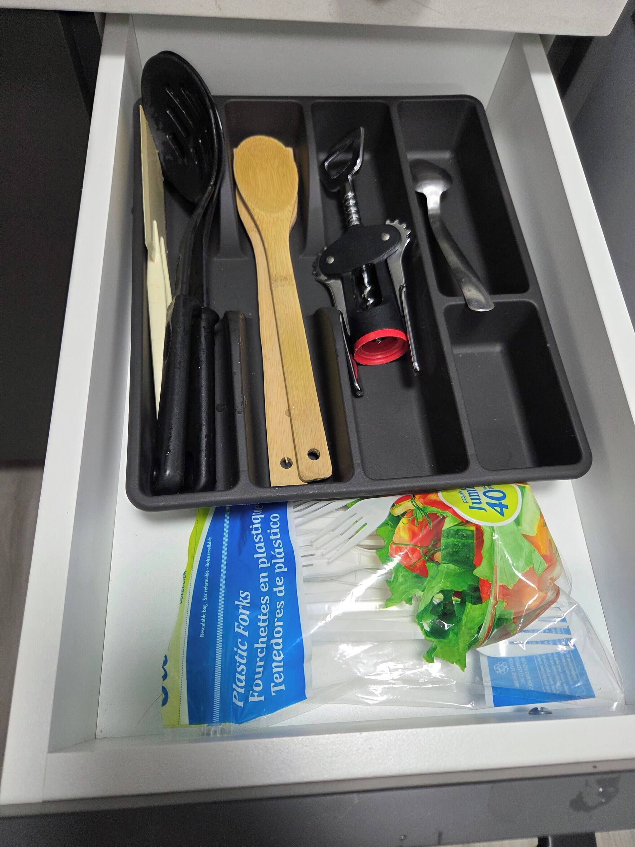 A drawer with a utensil set, a wooden spatula, a plastic bag of salad, and a blue label.