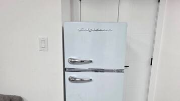 A white refrigerator with a handle and a control panel, located in a room with a wooden floor and a white door in the background.