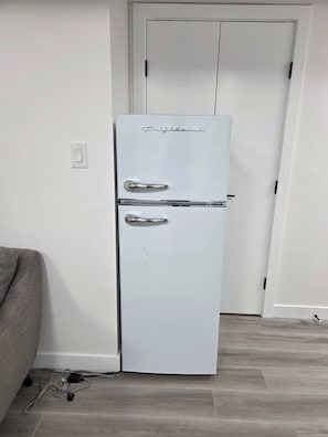 A white refrigerator with a handle and a control panel, located in a room with a wooden floor and a white door in the background. - The Cozy Corner Home (Winnipeg)
