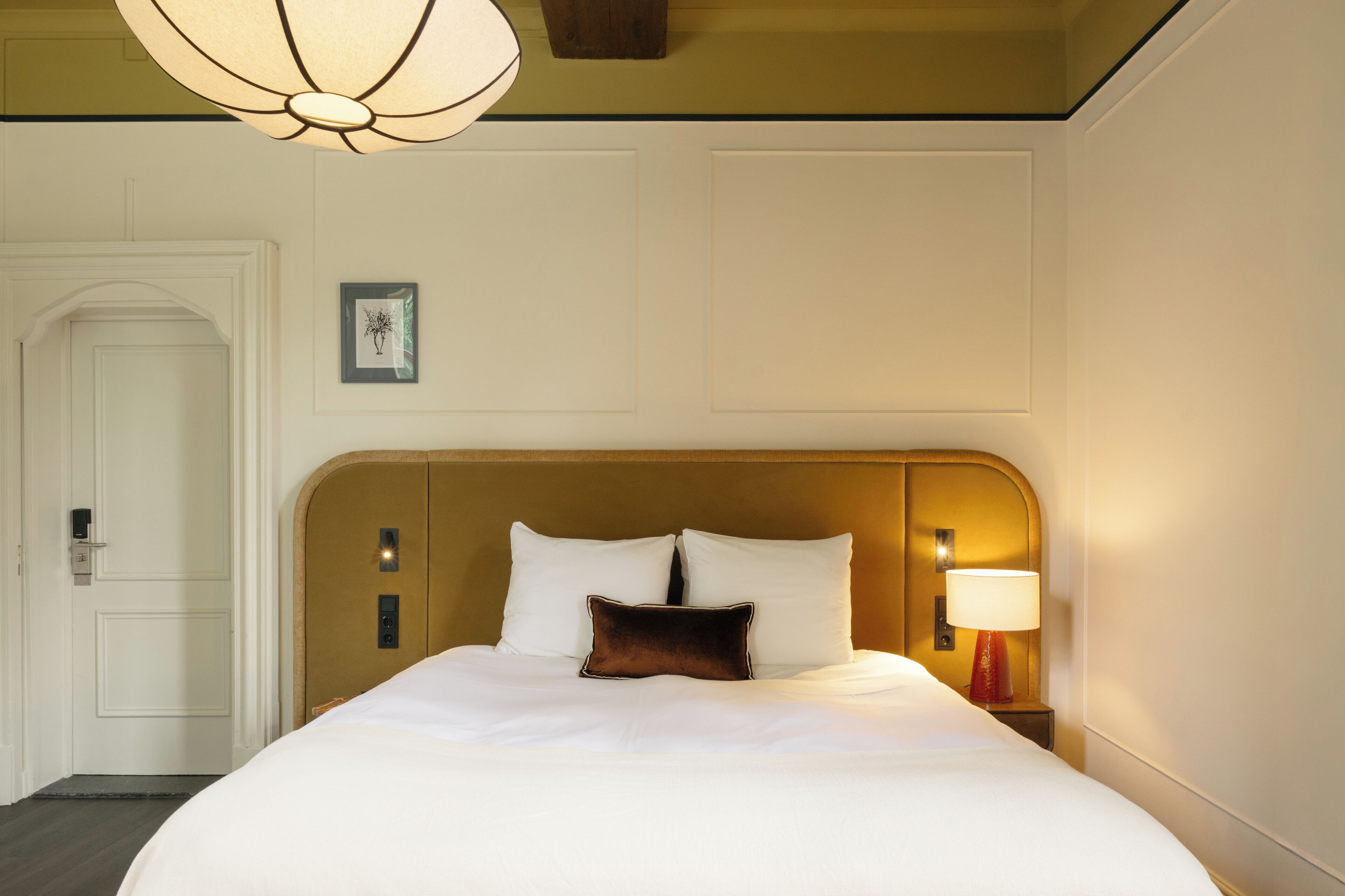 Habitación de hotel con una cama grande, una mesita de noche con lámpara, una puerta y un cuadro en la pared.