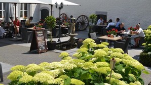 Un café al aire libre con mesas y sillas, rodeado de follaje y un edificio blanco.
