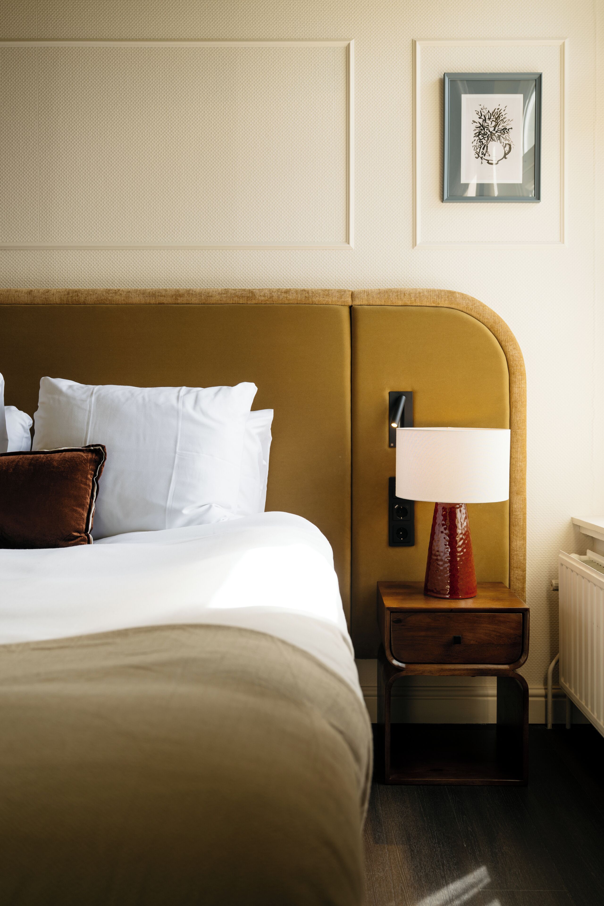 A neatly made bed with white linens and a brown headboard. There is a bedside table with a lamp and a framed picture on the wall above.