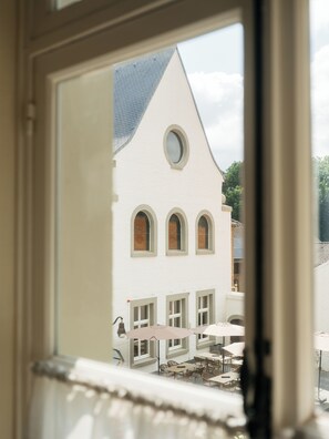 A view through a window of a building with a white facade, multiple windows, and a balcony area with tables and chairs.
