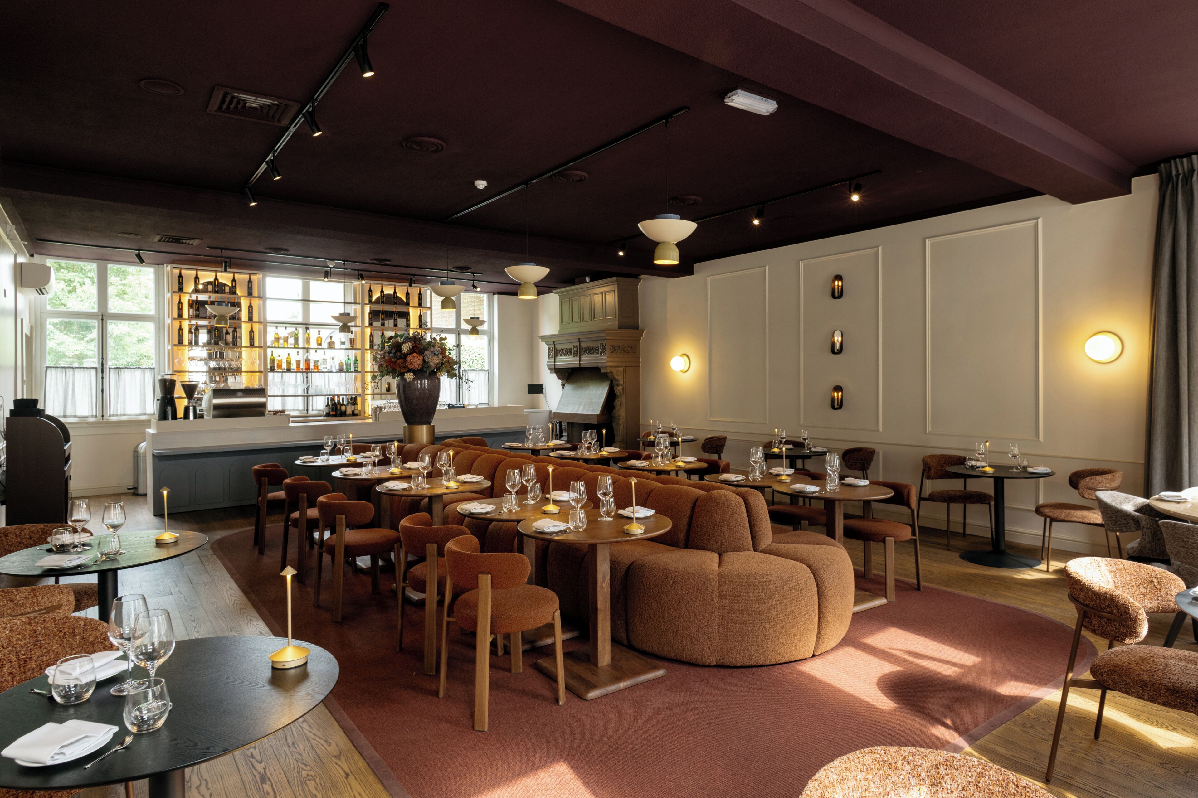 Een restaurant met ronde tafels, bruine beklede stoelen en een bar voorzien van flessenkasten.
