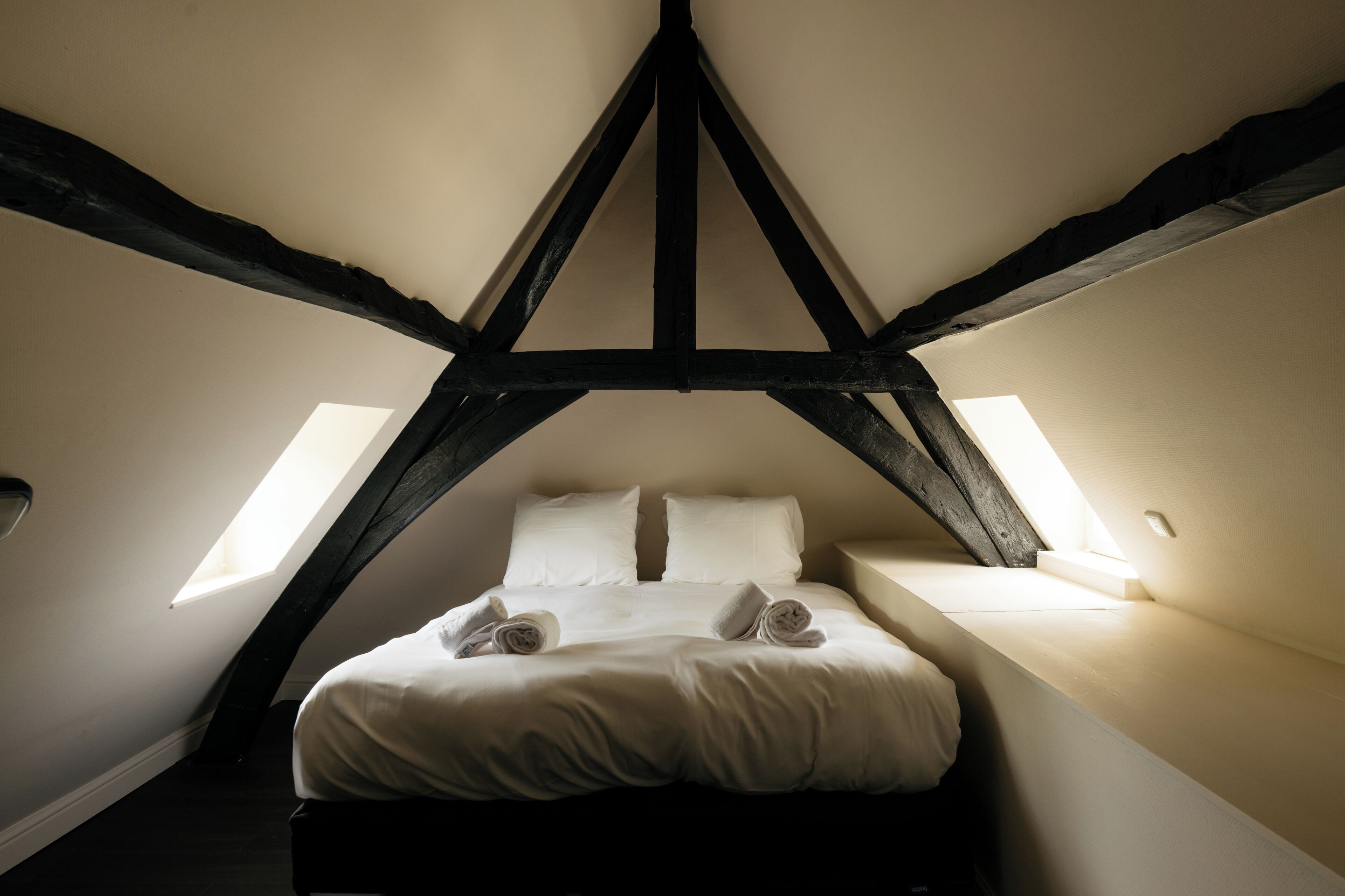 Un dormitorio con techo inclinado, vigas de madera a la vista, una cama con ropa blanca y dos tragaluces.