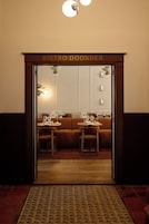Entrada de um restaurante com uma placa que diz 'BISTRO DOONDER'. No interior, mesas montadas com copos e talheres, e um sofá é visível ao fundo.