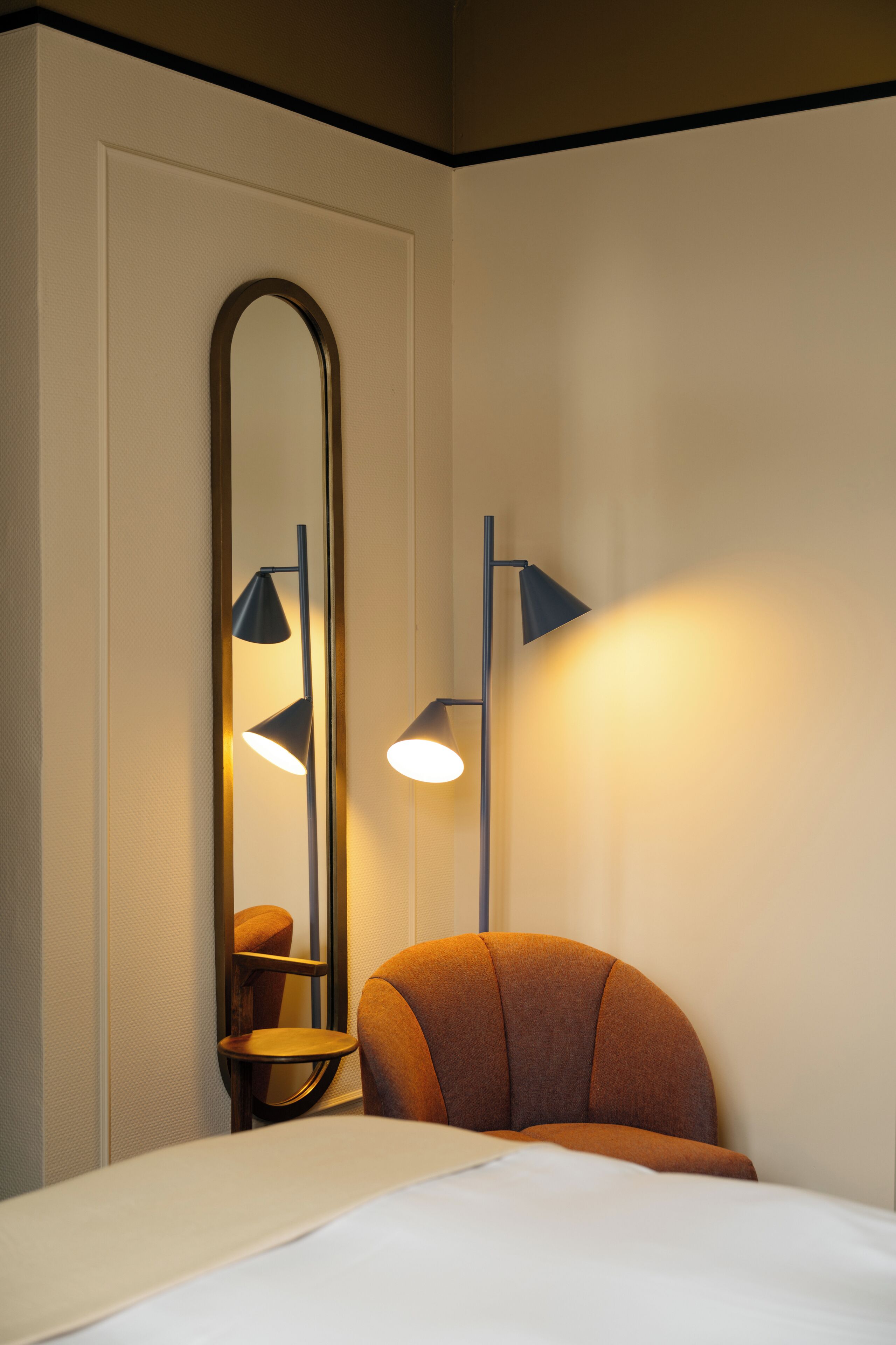 Una habitación de hotel con una cama, una silla, un espejo grande y dos lámparas fijadas a la pared.