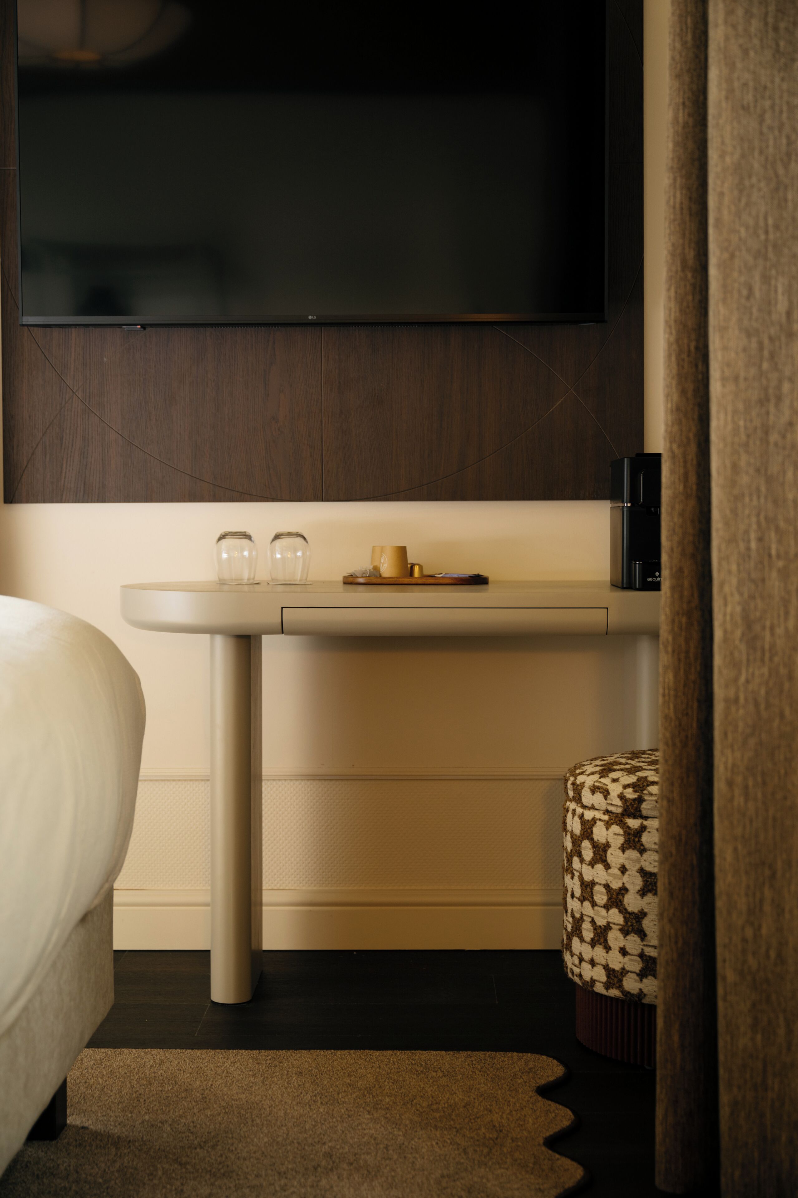 Una habitación de hotel moderna con una televisión de pantalla plana montada en la pared, una mesita de noche con lámpara, un taburete pequeño y una cama.