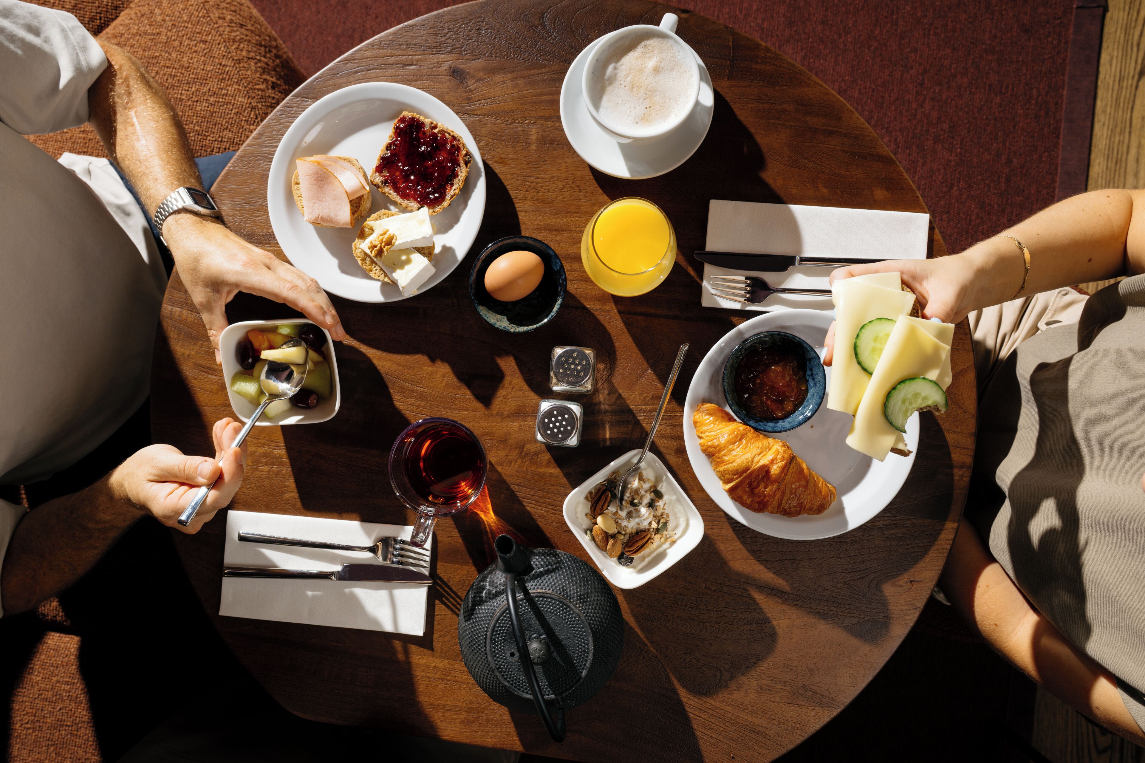 Een ronde houten tafel met diverse ontbijtartikelen, zoals toast, jam, eieren, een croissant en een glas sinaasappelsap.