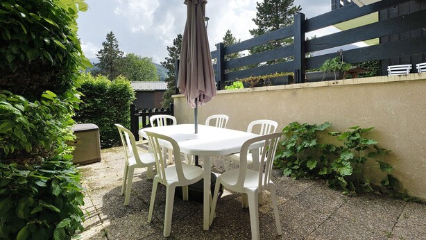 Un patio exterior con una mesa y sillas blancas, un paraguas morado y una cerca negra.