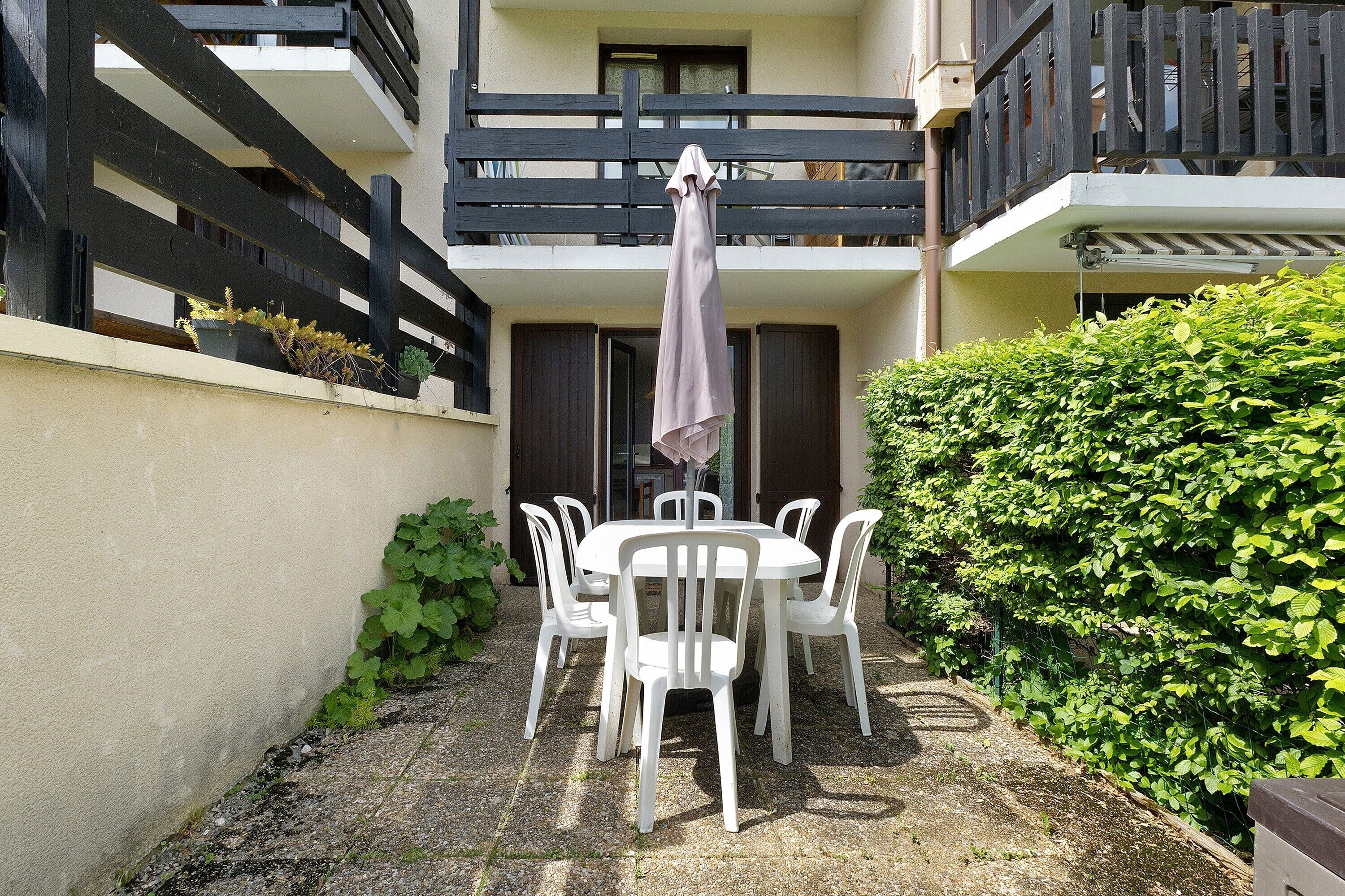 Une cour avec une table ronde et des chaises, un balcon avec une balustrade et une haie.