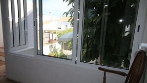 A room with a large window, a wooden chair, and a view of a garden and buildings outside. - CASA 8 - Sea View T3 (Luz)