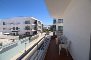 Ein Balkon mit weißen Stühlen, einem Tisch und einem Geländer mit Blick auf ein Wohngebiet mit weißen Gebäuden und blauem Himmel.