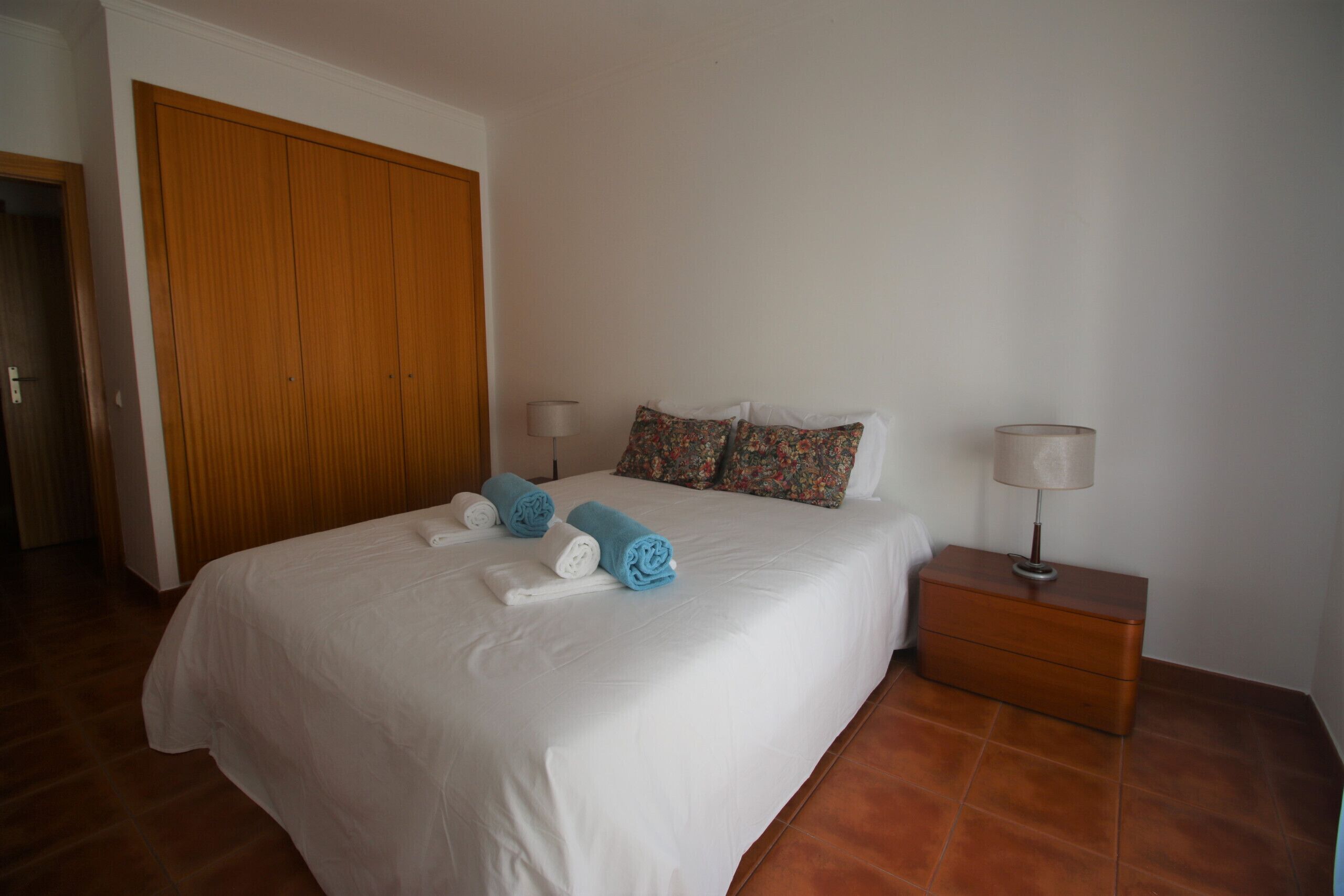 A bedroom with a bed, a wooden wardrobe, a nightstand with a lamp, and a tiled floor.
