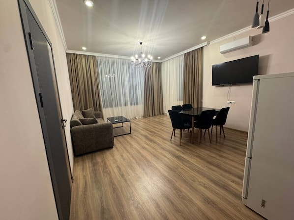 A modern living room with a sofa, a coffee table, a dining area with chairs, a chandelier, and a flat-screen TV mounted on the wall.