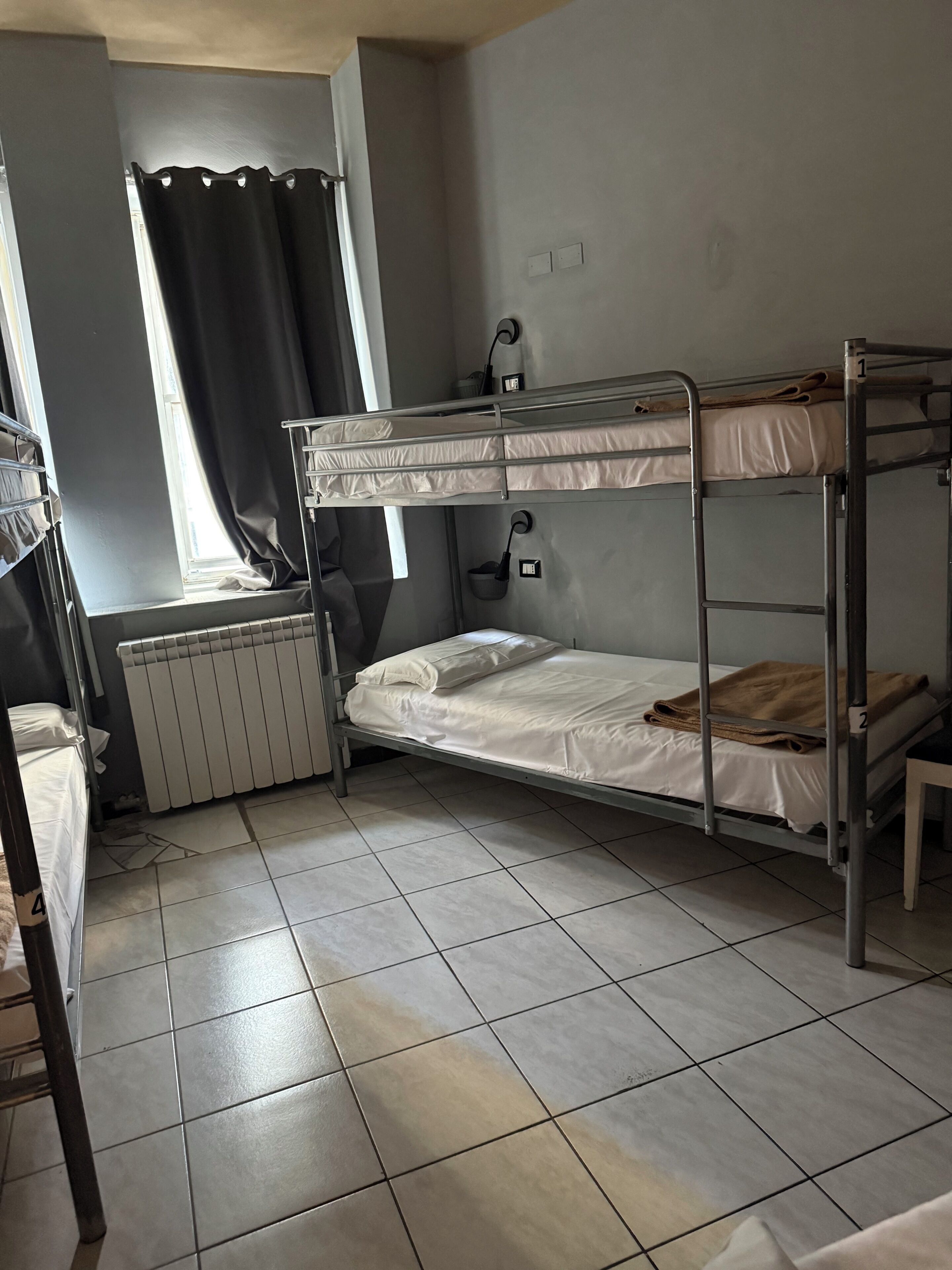 A bunk bed room with a radiator, a window with curtains, and a tiled floor.