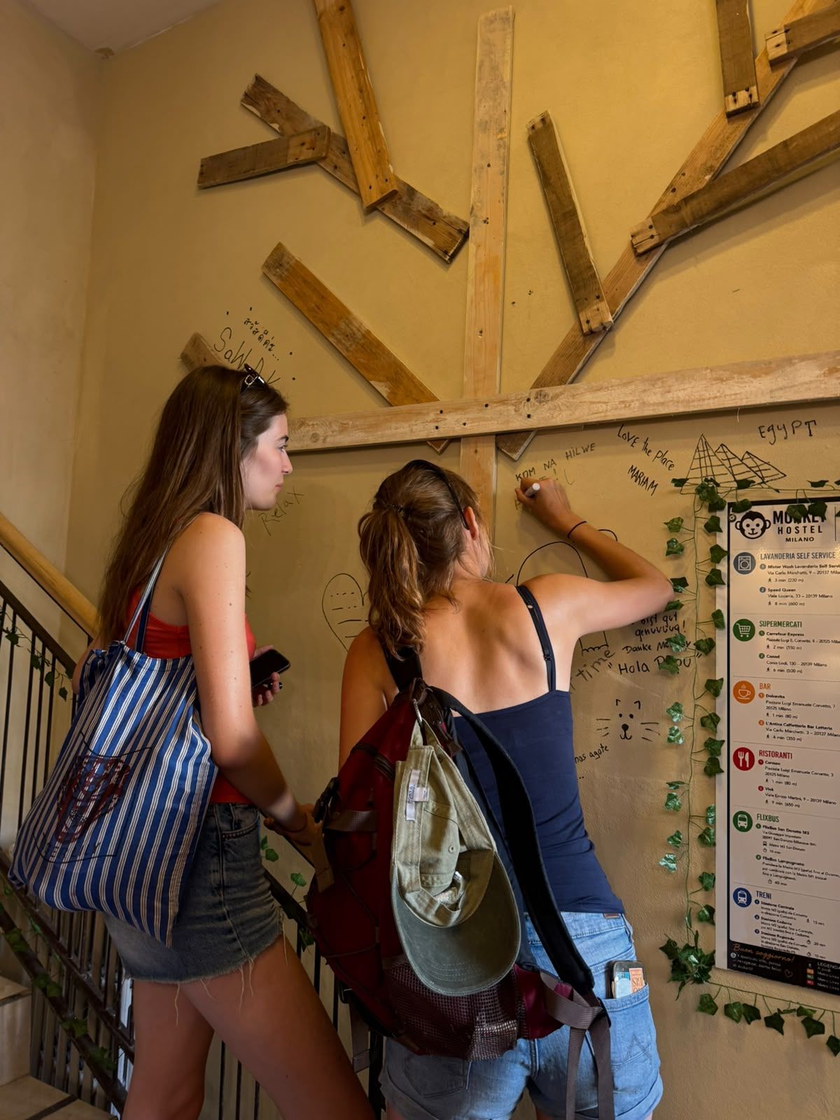 Dos personas observando una pared con diversos escritos y dibujos, incluyendo un mapa y una lista de servicios.