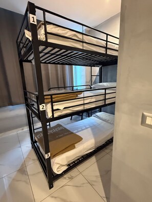 Cama litera con tres niveles, numerados del 1 al 3, en una habitación con suelo de baldosas y una ventana con cortinas.