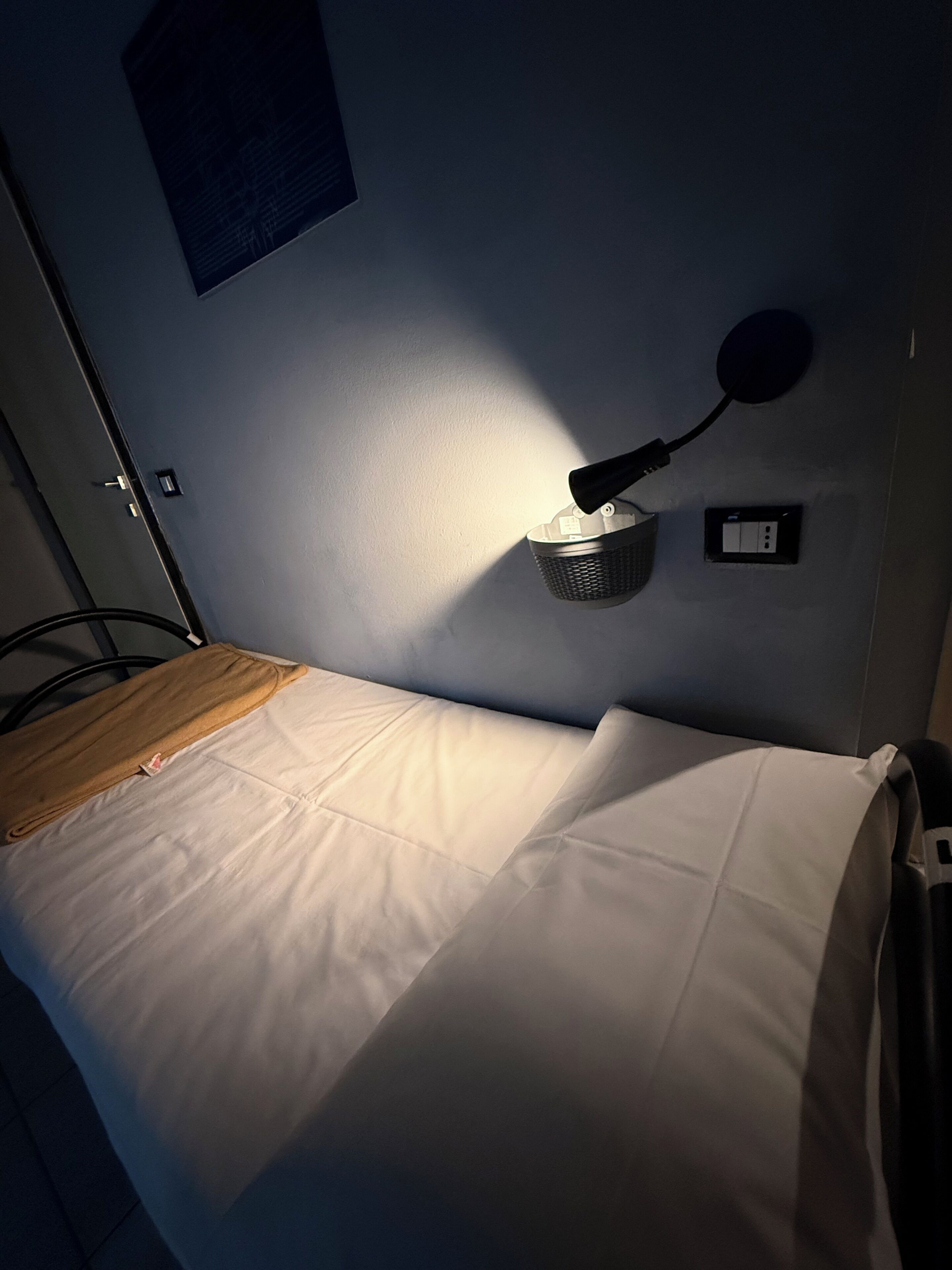 Un lit simple avec une literie blanche, une lampe fixée au mur et une prise de courant sur le mur.