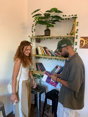 A person holding a book with a visible title, standing next to a bookshelf with books and a potted plant.