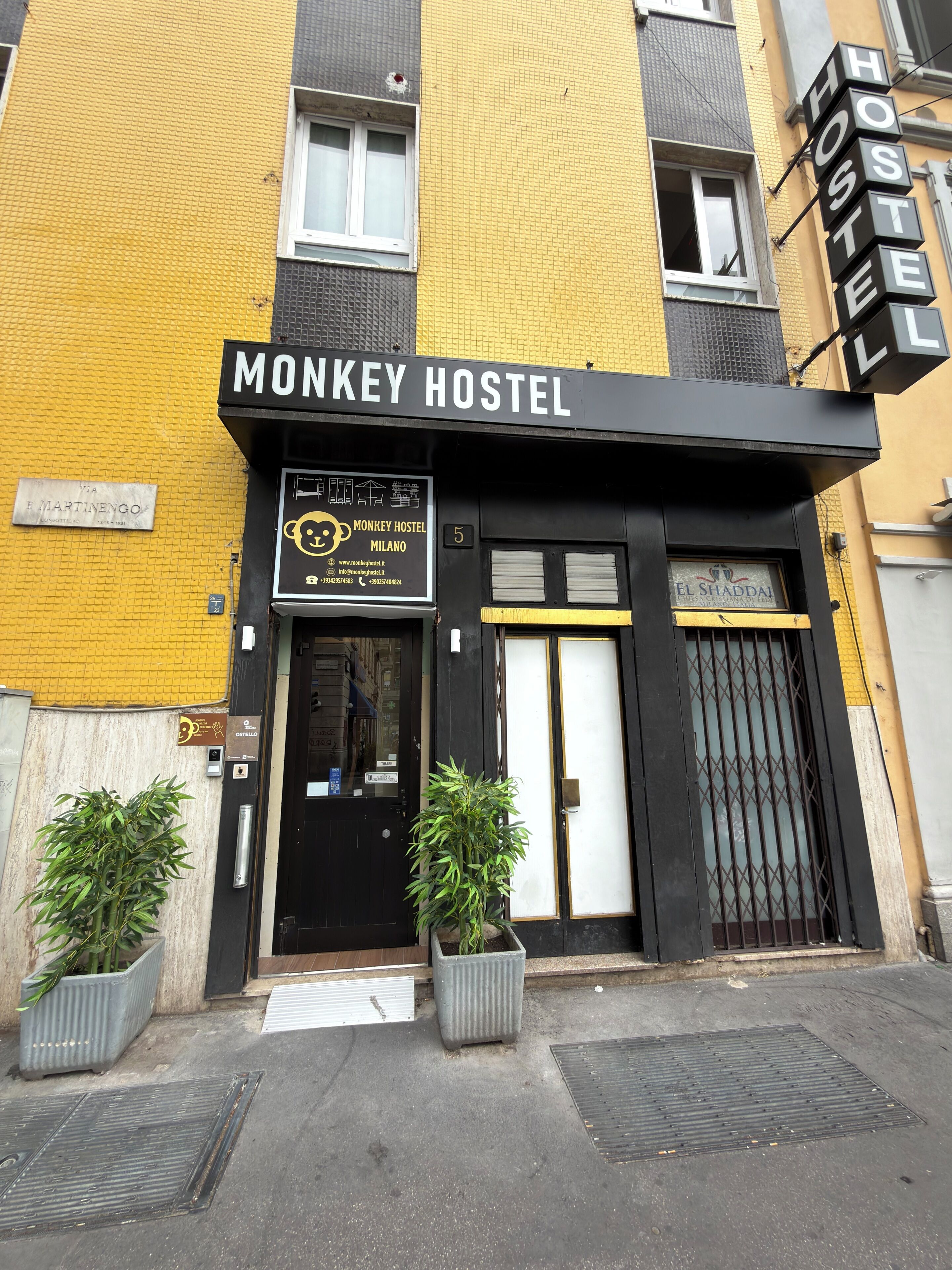 A building with a sign reading 'MONKEY HOSTEL MILANO' and a smaller sign for 'EL SHADAI'. There are two potted plants in front of the entrance.