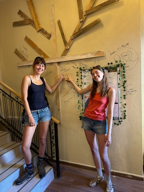 Zwei Personen stehen auf einer Treppe mit einer Holzkreuzkonstruktion und einer Wand mit verschiedenen Schriftzügen und Zeichnungen.