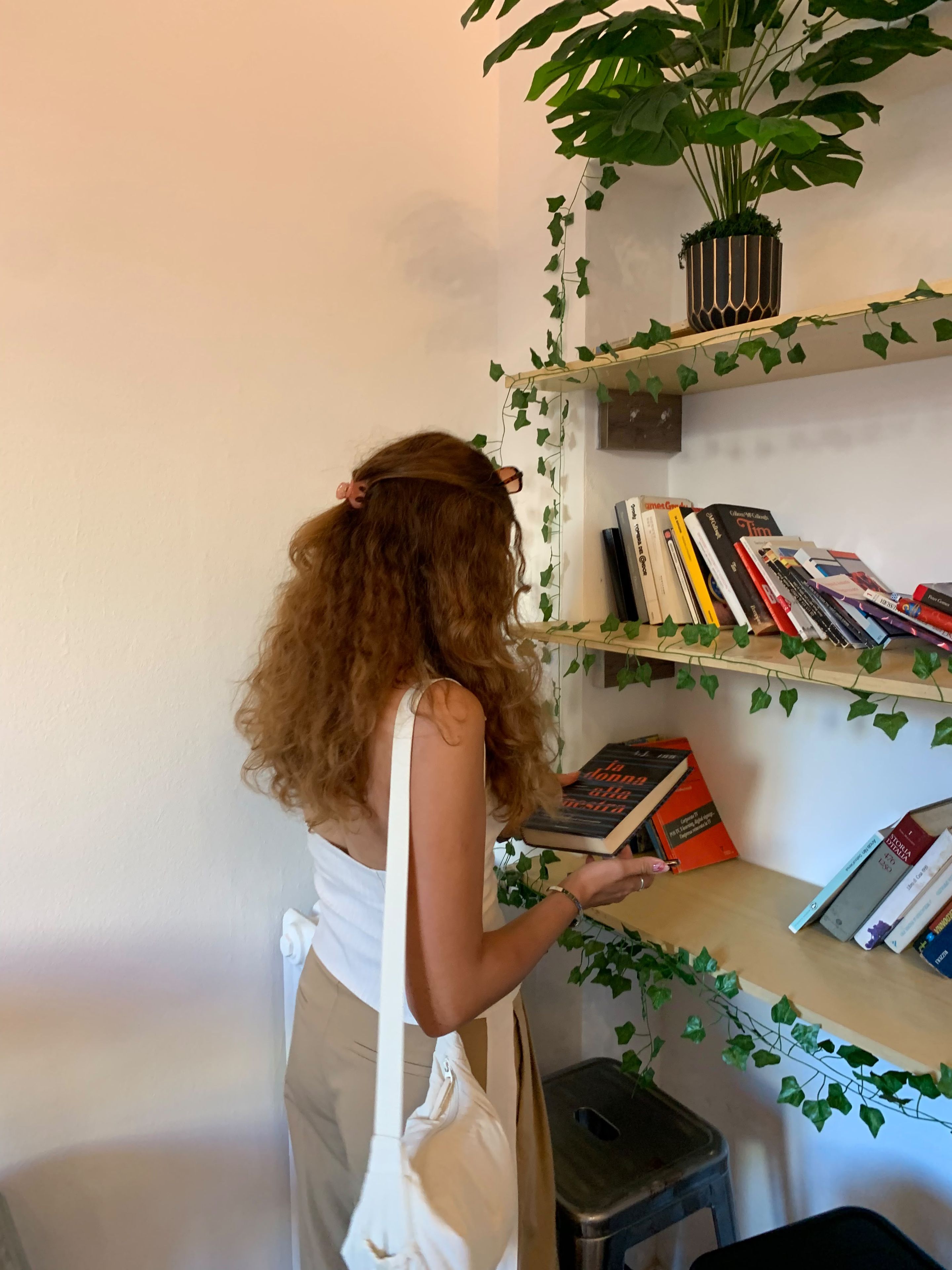 Eine Person liest ein Buch in einem Raum mit Bücherregal, Blumentopf und kleinem Hocker.