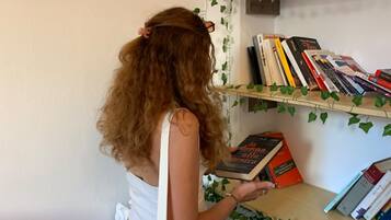 Eine Person liest ein Buch in einem Raum mit Bücherregal, Blumentopf und kleinem Hocker.