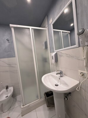 Un baño con un lavamanos blanco, un espejo, un secador de pelo de mano, una cabina de ducha de vidrio y un bidé.