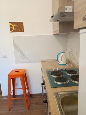 A compact kitchen with a gas stove, a kettle, and a small stool.