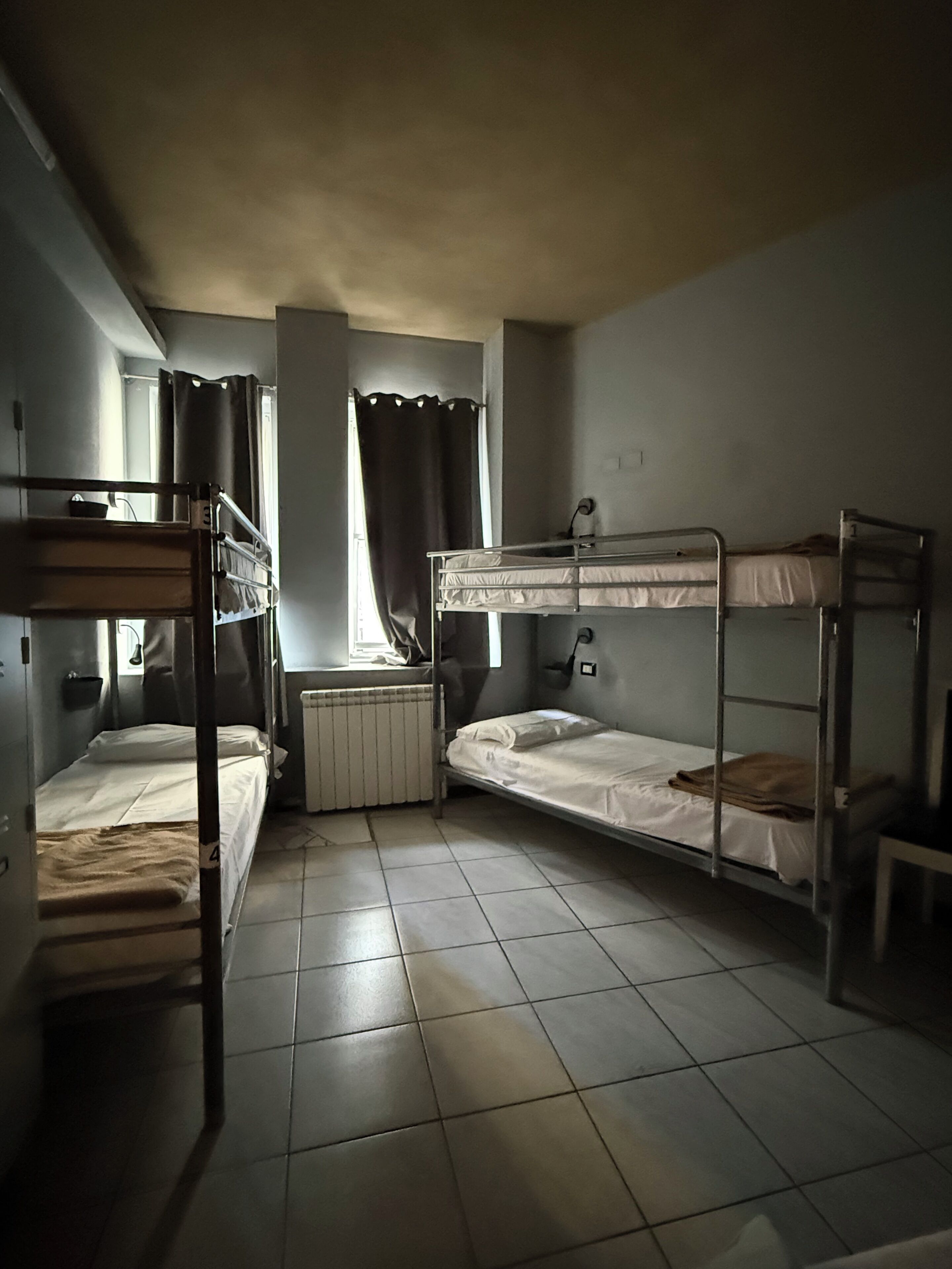 Une chambre de dortoir avec des lits superposés, un radiateur, une fenêtre avec des rideaux et un sol carrelé.
