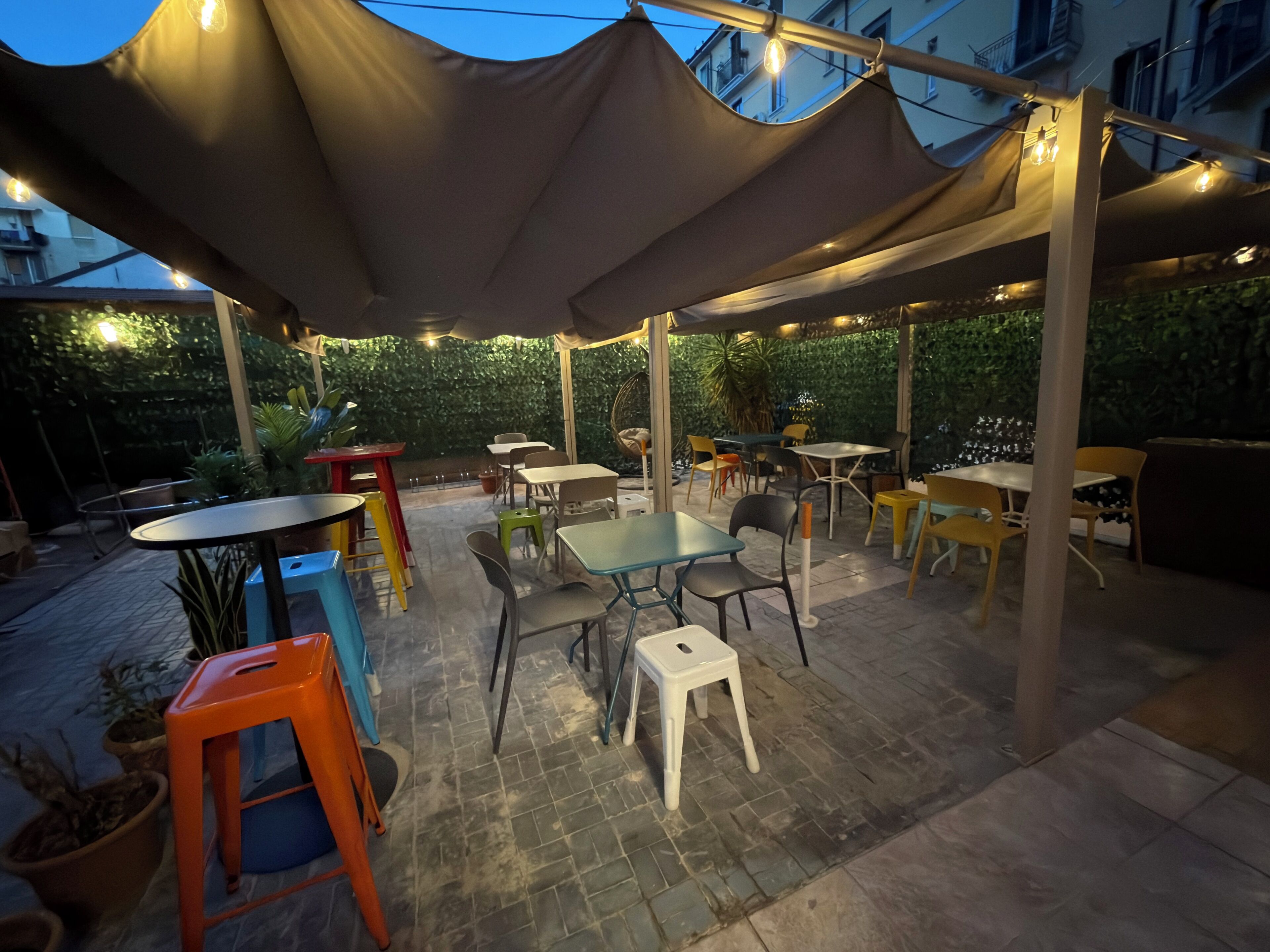 Un patio al aire libre con mesas y sillas, rodeado de follaje y un edificio al fondo.