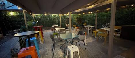 An outdoor patio area with tables and chairs, surrounded by greenery and a building in the background.