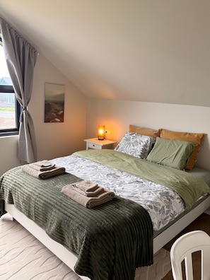 A neatly made bed with a patterned bedspread, a green blanket, and folded towels. There is a bedside lamp, a nightstand, and a chair. - Charming Holiday House in Álftarnes Iceland With Scenic Views (Garðabær)