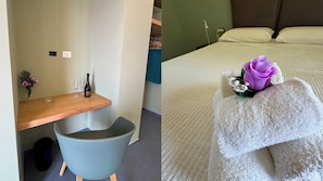 Ein Hotelzimmer mit einem Holztisch, einem Stuhl, einer kleinen Mülltonne, einem Bett mit weißer Decke und einer violetten Blume auf dem Bett.