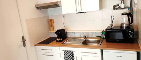 Cocina compacta con armarios blancos, un lavamanos de acero inoxidable, cafetera, microondas y hervidor.