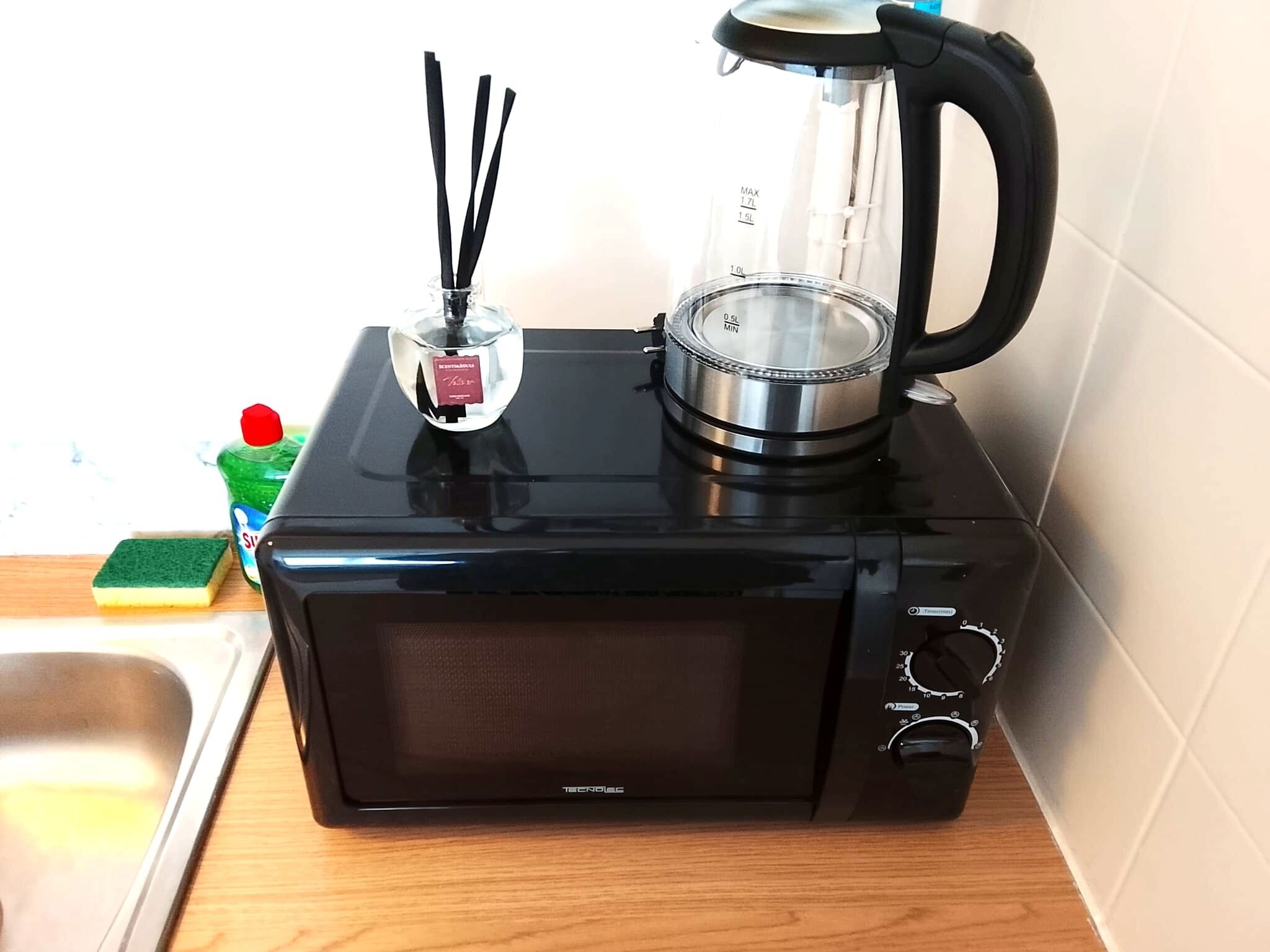 Een zwarte magnetronoven met een transparante glazen bak, een spons en een fles afwasmiddel op een houten aanrecht.