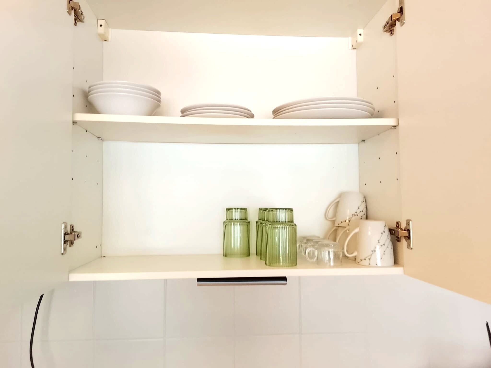 Un pensile da cucina con ripiani bianchi, contenente piatti, tazze e barattoli di vetro verde.