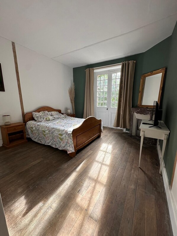 A bedroom with a wooden bed, a bedside table, a desk with a chair, a mirror, and a window with curtains.