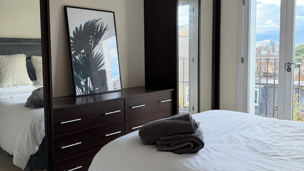 A bedroom with a large bed, a dark wooden dresser, a large mirror, and a city view through a sliding door. - Spacious two bedroom, split level flat in the heart of Wandsworth. (London)