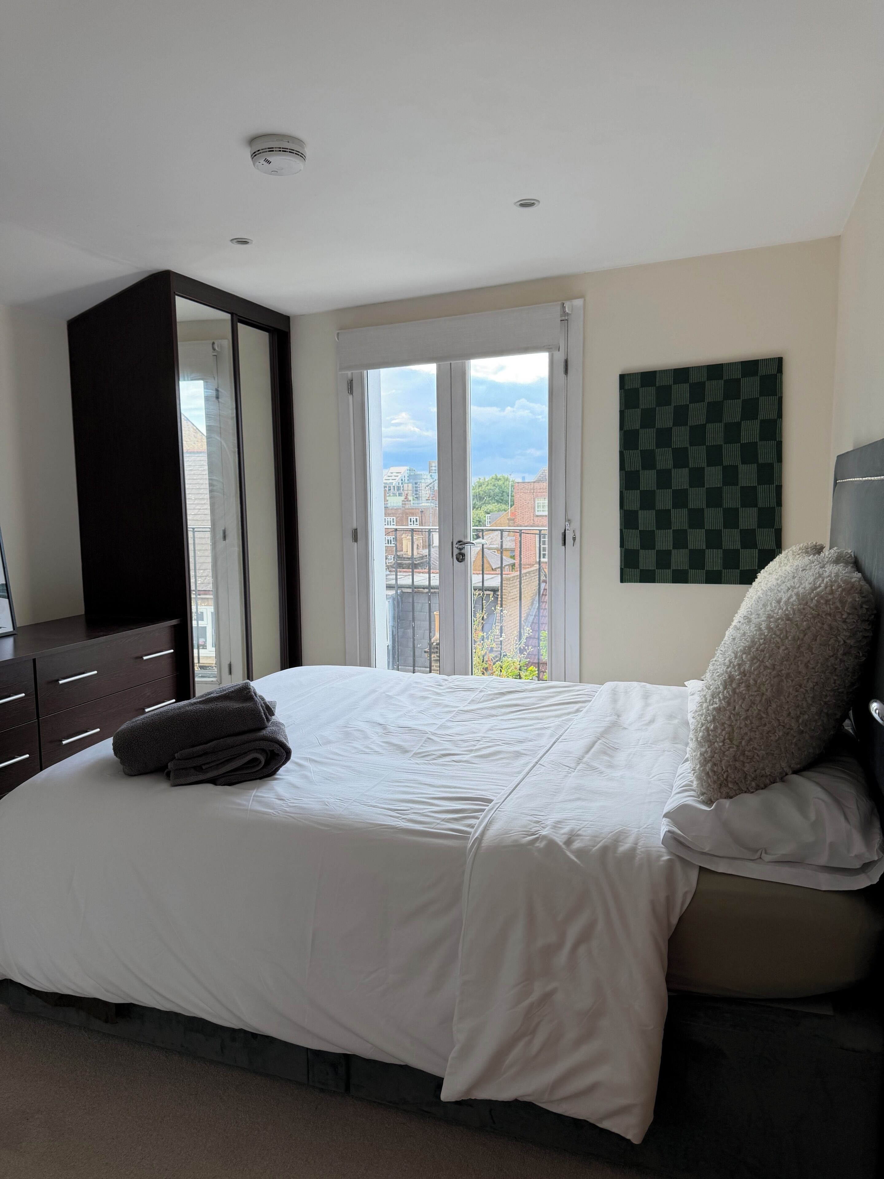 A bedroom with a bed, a wardrobe, a checkered wall art, and a balcony with a view.
