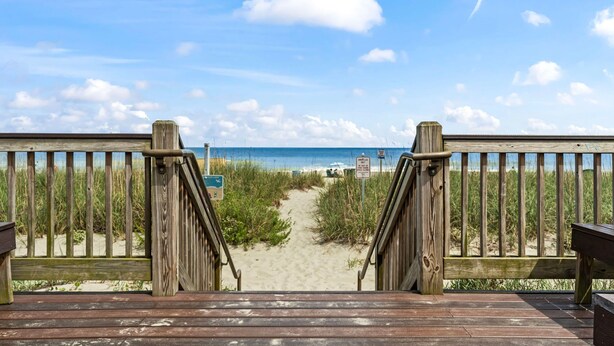 Een houten terras dat naar een strand leidt, met een heldere lucht.
