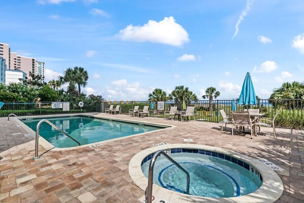 A swimming pool with a hot tub, lounge chairs, and a view of palm trees and buildings. - The Ocean Escape | Prime Myrtle Beach Getaway (Myrtle Beach)