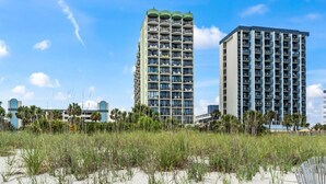 Een hoog hotel met meerdere balkons, een strand op de voorgrond en heldere luchten.