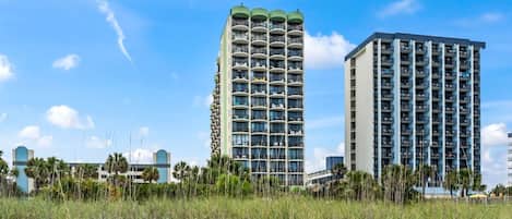 A high-rise hotel with multiple balconies, a beach in the foreground, and clear skies.