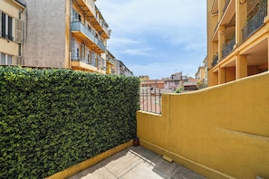 Ein schmaler Gang mit einer Hecke, einer gelben Wand und einem Balkon mit Metallgitter.