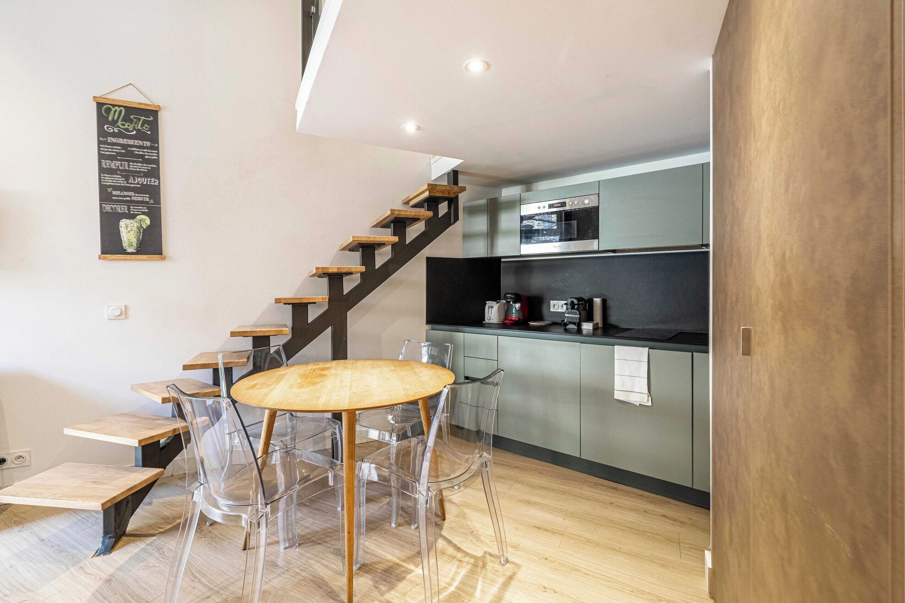 A modern kitchen with a built-in oven, a coffee machine, and a small dining area with a round table and clear chairs.