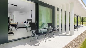 Uma área de estar moderna ao ar livre, com mesas e cadeiras de vidro, um alpendre envidraçado e vista para o espaço interior.